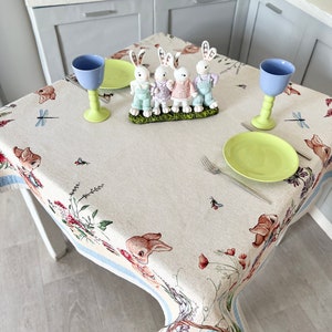 Puede incluir: Un mantel blanco con un borde azul presenta un patr&oacute;n floral con conejos, lib&eacute;lulas y una cesta de huevos de Pascua. Dos vasos azules y un plato verde est&aacute;n colocados en la mesa con un conjunto de cuatro figuras de conejo blanco.