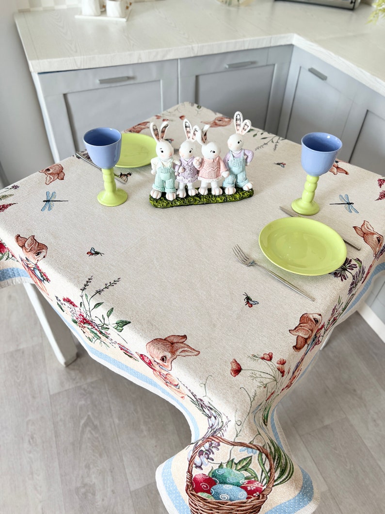 Puede incluir: Un mantel blanco con un borde azul presenta un patr&oacute;n floral con conejos, lib&eacute;lulas y una cesta de huevos de Pascua. Dos vasos azules y un plato verde est&aacute;n colocados en la mesa con un conjunto de cuatro figuras de conejo blanco.