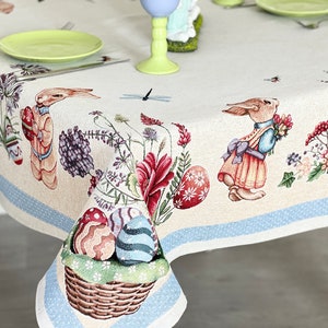 Puede incluir: Un mantel blanco con un borde azul y un patr&oacute;n repetido de conejos de Pascua, flores y huevos. El mantel est&aacute; decorado con una cesta de huevos de Pascua coloridos.