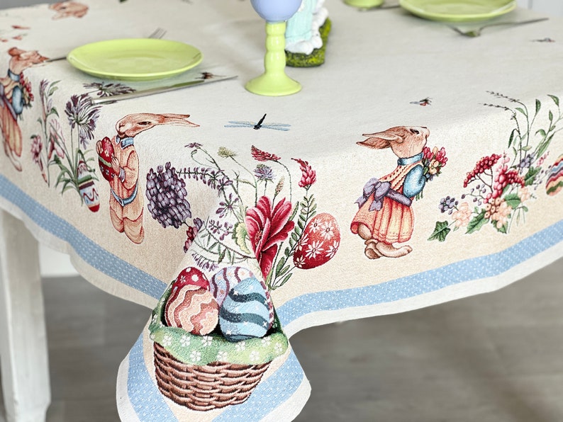 Puede incluir: Un mantel blanco con un borde azul y un patr&oacute;n repetido de conejos de Pascua, flores y huevos. El mantel est&aacute; decorado con una cesta de huevos de Pascua coloridos.