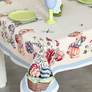 Puede incluir: Un mantel blanco con un borde azul y un patr&oacute;n floral con conejos, huevos y una lib&eacute;lula. El mantel est&aacute; puesto con un plato verde, un tenedor de plata y un vaso azul claro.