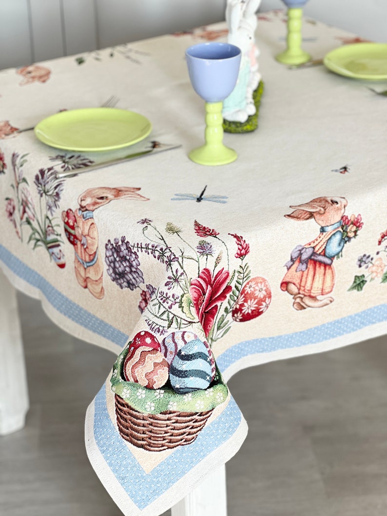 Puede incluir: Un mantel blanco con un borde azul y un patr&oacute;n floral con conejos, huevos y una lib&eacute;lula. El mantel est&aacute; puesto con un plato verde, un tenedor de plata y un vaso azul claro.