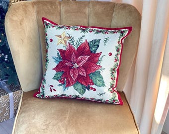 Op de afbeelding: Vierkant decoratief kussen met een kerststerontwerp. Het kussen heeft een rode rand en een witte achtergrond met een grote rode kerststerbloem, groene bladeren, bessen en een gouden ster. Het ligt op een gouden stoel.