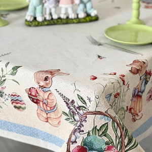 Puede incluir: Un mantel beige con un borde azul presenta un tema de Pascua con conejos, flores y huevos. Una cesta llena de huevos coloridos se encuentra en la esquina inferior derecha.
