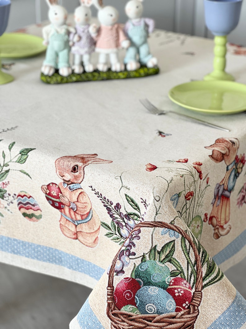 Puede incluir: Un mantel beige con un borde azul presenta un tema de Pascua con conejos, flores y huevos. Una cesta llena de huevos coloridos se encuentra en la esquina inferior derecha.