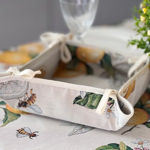 May include: A white fabric bread basket with a lemon and bee design. The basket is folded and has white ties.