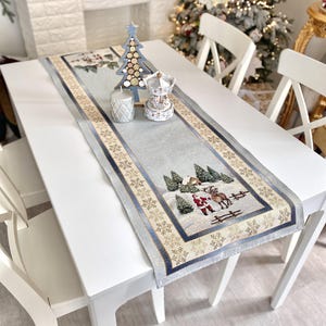 Può includere: Un tavolo bianco con un runner da tavola natalizio blu e oro che presenta una scena invernale con una casa, alberi e Babbo Natale. Il runner ha un motivo a fiocco di neve e un bordo dorato e blu. C'è un albero di Natale in legno e una giostra bianca sul tavolo.