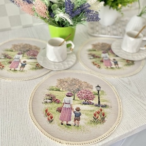 Puede incluir: Juego de cuatro manteles individuales redondos con una ilustración de estilo vintage de una mujer y un niño en un parque. Cada mantel individual tiene un borde color crema y una escena detallada con árboles, flores y una farola. Tazas y platillos blancos encima.