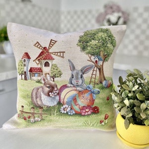 May include: A decorative pillow with a white background featuring a colorful illustration of two bunnies holding Easter eggs, a windmill, and a tree. The pillow is perfect for adding a touch of spring to your home decor.