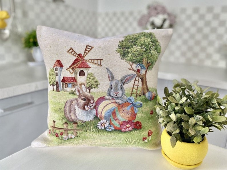 May include: A decorative pillow with a white background featuring a colorful illustration of two bunnies holding Easter eggs, a windmill, and a tree. The pillow is perfect for adding a touch of spring to your home decor.