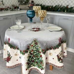 Peut inclure: Une nappe ronde blanche avec un motif d'arbre de Noël. La nappe présente un sapin de Noël vert avec des décorations rouges et dorées, entouré d'une guirlande de verdure et de baies rouges. La nappe est dressée avec deux couverts, chacun avec une assiette blanche et un verre transparent.