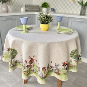 May include: A round, white tablecloth with a whimsical Easter-themed design. The tablecloth features a border with illustrations of bunnies, houses, and a windmill. The tablecloth is set on a table with two plates and glasses.