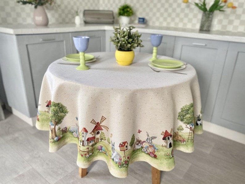 May include: A round, white tablecloth with a whimsical Easter-themed design. The tablecloth features a border with illustrations of bunnies, houses, and a windmill. The tablecloth is set on a table with two plates and glasses.