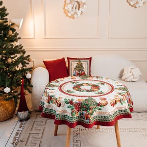 May include: A round white tablecloth with a red border features a festive Christmas design with a wreath, ornaments, and presents. The tablecloth is on a round wooden table with wooden legs.