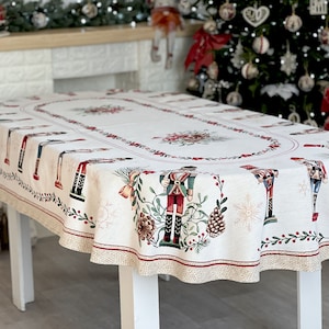 May include: A white oval tablecloth with a festive Christmas design featuring nutcracker soldiers, pine cones, holly, and snowflakes. The tablecloth has a beige and red border.