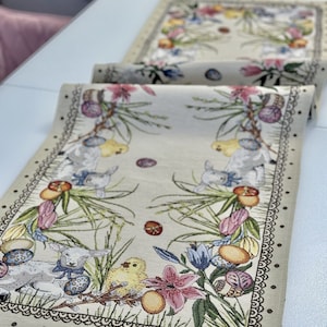 May include: A beige table runner with an embroidered Easter design. The design features chicks, lambs, flowers, and eggs. The border is a brown scalloped edge with black dots.