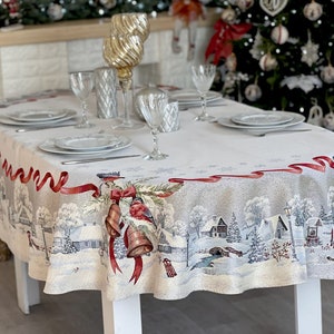 Puede incluir: Un mantel ovalado blanco con una escena de un pueblo de invierno impresa en él. El mantel tiene un borde de cinta roja con un patrón repetido de copos de nieve. El mantel está puesto con cuatro cubiertos, cada uno con un plato blanco, un vaso y cubiertos.