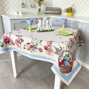 Puede incluir: Un mantel blanco con un borde azul presenta un dise&ntilde;o de Pascua colorido con conejos, flores y huevos. El mantel est&aacute; puesto sobre una mesa con patas blancas y est&aacute; decorado con vasos azules, un plato de huevos coloridos y una cesta de huevos.