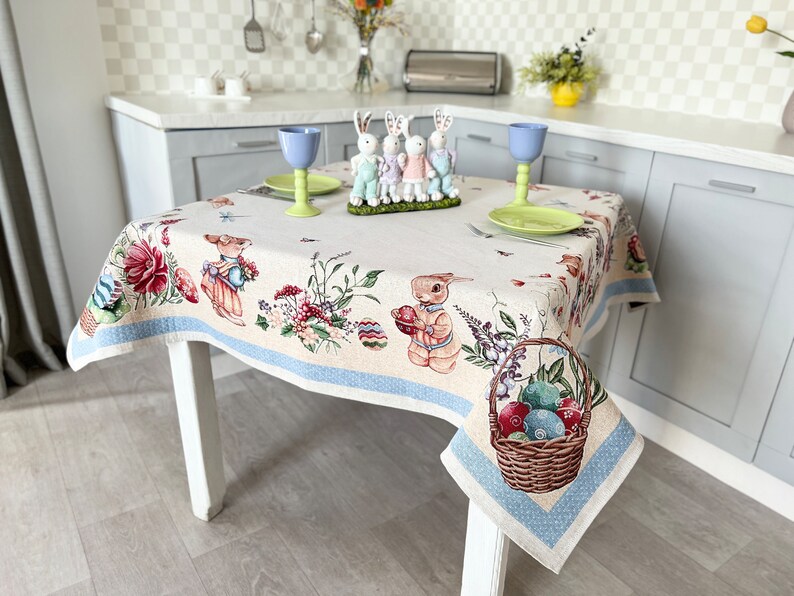 Puede incluir: Un mantel blanco con un borde azul presenta un dise&ntilde;o de Pascua colorido con conejos, flores y huevos. El mantel est&aacute; puesto sobre una mesa con patas blancas y est&aacute; decorado con vasos azules, un plato de huevos coloridos y una cesta de huevos.