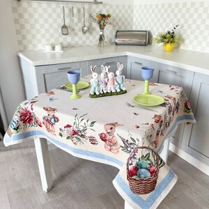 Puede incluir: Un mantel blanco con un borde azul presenta un patr&oacute;n floral con conejos y huevos de Pascua. El mantel est&aacute; puesto sobre una mesa blanca con una cesta de huevos de Pascua coloridos.
