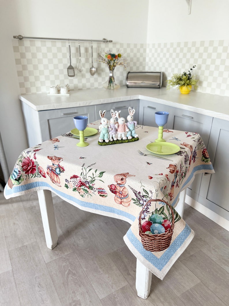 Puede incluir: Un mantel blanco con un borde azul presenta un patr&oacute;n floral con conejos y huevos de Pascua. El mantel est&aacute; puesto sobre una mesa blanca con una cesta de huevos de Pascua coloridos.
