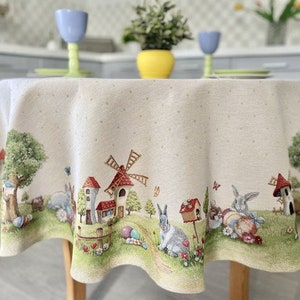 May include: A round, white tablecloth with a whimsical Easter scene. The tablecloth features a windmill, houses, bunnies, and eggs. The tablecloth has a subtle polka dot pattern.
