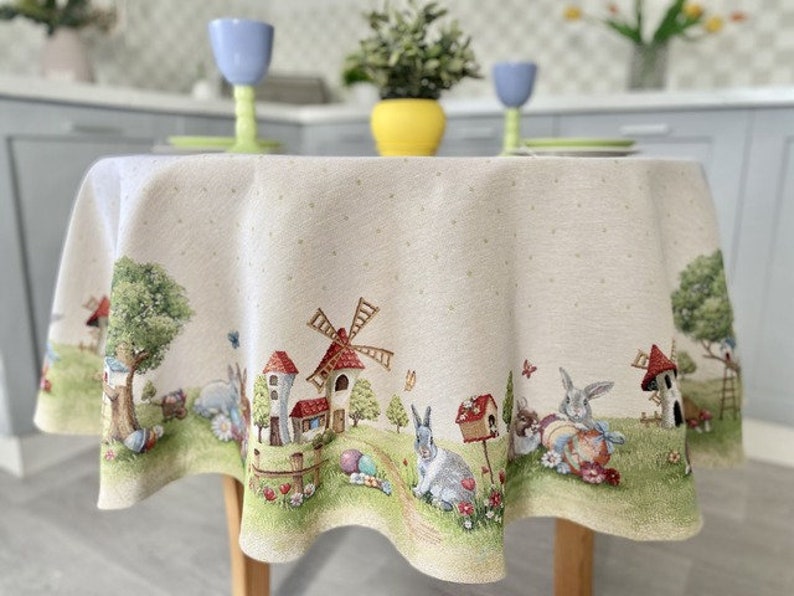 May include: A round, white tablecloth with a whimsical Easter scene. The tablecloth features a windmill, houses, bunnies, and eggs. The tablecloth has a subtle polka dot pattern.