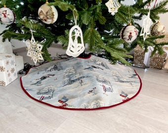 May include: A white and gray Christmas tree skirt with a winter scene design. The skirt has a red border and is decorated with small houses, trees, and skiers. The skirt is placed under a Christmas tree with ornaments and lights.