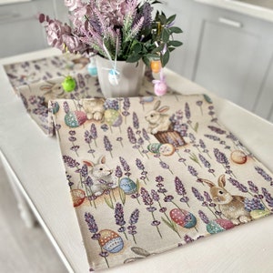 Op de afbeelding: Een beige tafelkleed met een lavendel- en konijnenontwerp. De tafelkleed heeft een herhalend patroon van lavendelbloemen, paaseieren en konijnen. De tafelkleed is perfect om een vleugje lente aan uw tafel toe te voegen.