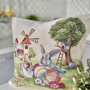 May include: A decorative pillow featuring a whimsical Easter scene with two bunnies, a windmill, a treehouse, and colorful Easter eggs. The pillow is made of a beige fabric with a tapestry-like texture.