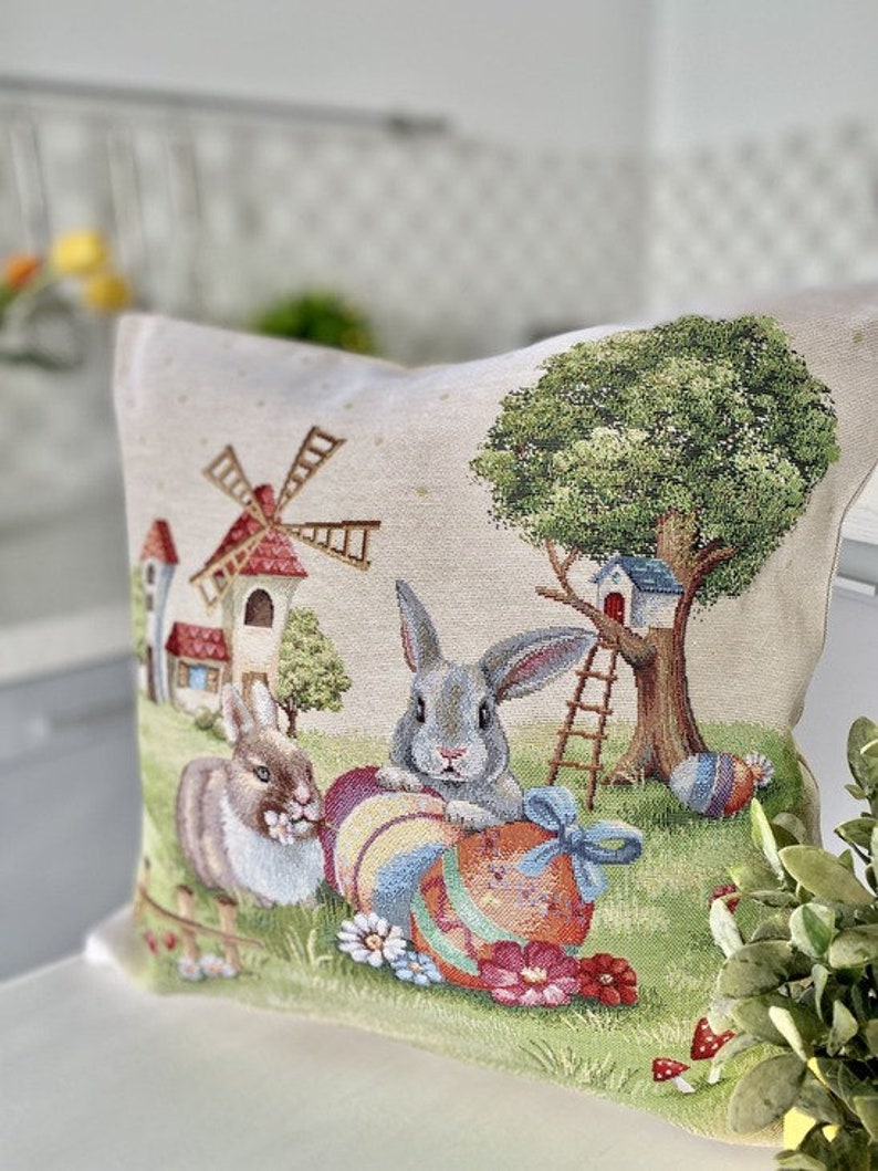 May include: A decorative pillow featuring a whimsical Easter scene with two bunnies, a windmill, a treehouse, and colorful Easter eggs. The pillow is made of a beige fabric with a tapestry-like texture.