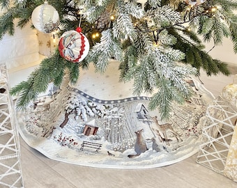 May include: A white Christmas tree skirt with a winter scene featuring a house, trees, and animals. The skirt has a white background with a blue and brown design. The skirt is decorated with snowflakes and stars.