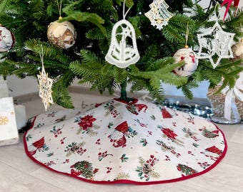 May include: A white and red Christmas tree skirt with a gnome pattern. The skirt has a red border and is decorated with holly berries and pine cones.