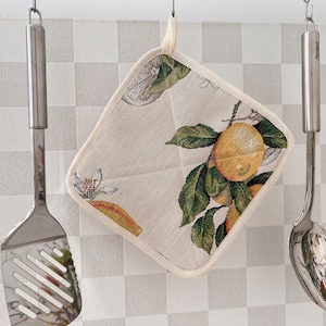 Puede incluir: Un agarrador de olla blanco con un diseño de impresión de limón. El agarrador de olla está colgado de un gancho con otros utensilios de cocina.