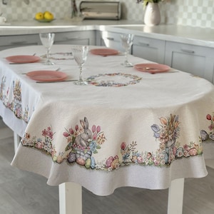 May include: A white oval tablecloth with a gray border features a floral design with bunnies and eggs. The tablecloth is laid on a table with four place settings.