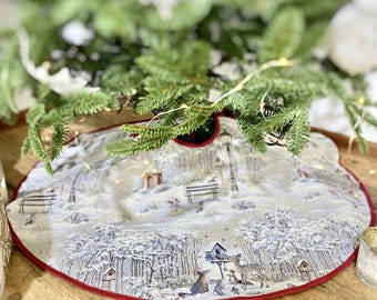 May include: A white and beige Christmas tree skirt with a winter scene design. The skirt features a red border and a snowy forest with animals and a birdhouse.