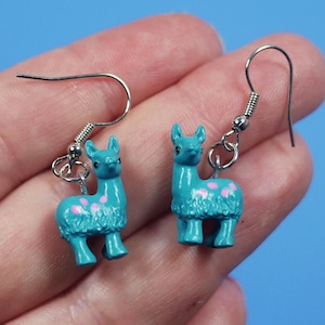 May include: Teal llama dangle earrings with pink accents. The earrings have silver hooks.
