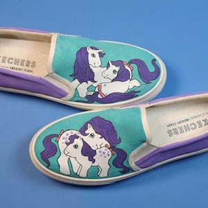 Könnte beinhalten: Paar Skechers-Schlupfschuhe mit türkis-violettem Design. Jeder Schuh zeigt eine weiße Pony-Grafik mit lila Haaren und einer roten Schleife. Die Schuhe haben eine weiße Sohle und das Wort "Skechers" ist innen aufgedruckt.