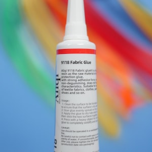 May include: A white plastic squeeze tube of 9118 Fabric Glue with a pointed applicator tip. The label features black text detailing product usage and safety instructions. The tube is positioned vertically against a blurred, colorful background.