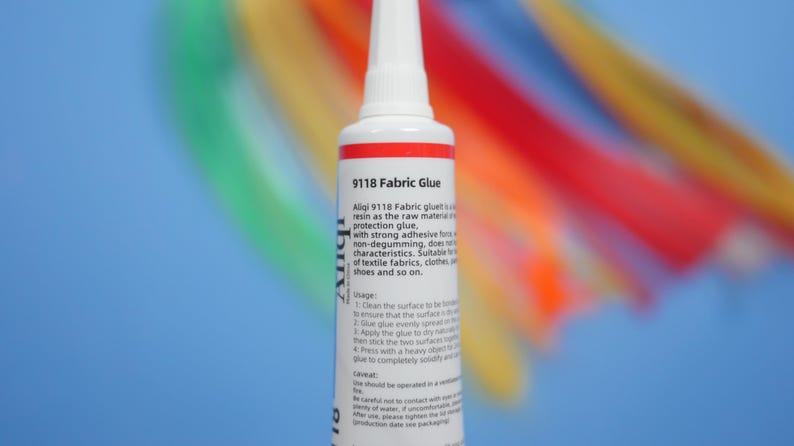 May include: A white plastic squeeze tube of 9118 Fabric Glue with a pointed applicator tip. The label features black text detailing product usage and safety instructions. The tube is positioned vertically against a blurred, colorful background.