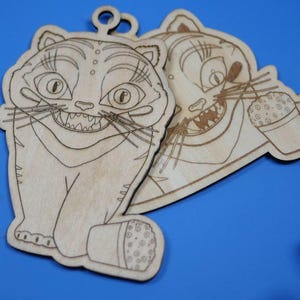 May include: Two wooden cat-shaped ornaments with engraved details. One cat is sitting, the other is holding a drink. Both have large grins and visible teeth. The ornaments are light brown and have a loop for hanging.