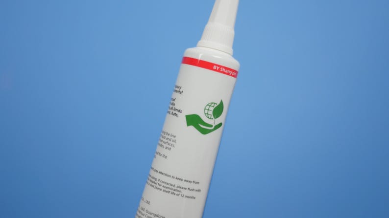May include: A white cylindrical adhesive tube with a pointed nozzle cap. The label features a red horizontal stripe at the top with the text BY Shang pin, a green icon of a hand holding a globe with a leaf, and black printed instructional text.