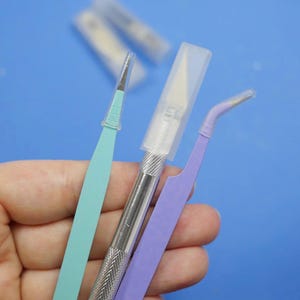 May include: A collection of craft tools, including a light blue precision knife, a silver craft knife with a protective cap, and purple tweezers. The tools are held against a blue background, suggesting a crafting or hobby theme.