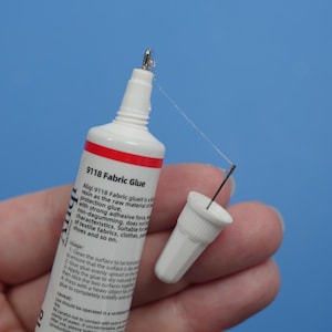 May include: A white and red tube of 9118 Fabric Glue with a pointed applicator tip and a detached white cap. The tube features printed text detailing usage instructions and product information, with a thin strand of adhesive visible at the tip.