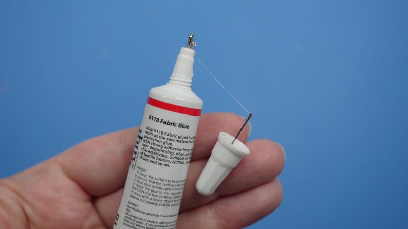 May include: A white and red tube of 9118 Fabric Glue with a pointed applicator tip and a detached white cap. The tube features printed text detailing usage instructions and product information, with a thin strand of adhesive visible at the tip.