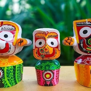 May include: Three hand-painted wooden figurines depicting a deity with large eyes, a red mouth, and a white face. The figurines are painted in bright colors, including red, yellow, green, and blue. The figurines are standing on a wooden surface.