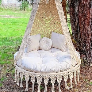 Könnte beinhalten: Ein weißer Makramee-Hängesessel mit einem runden Kissen und zwei Kissen. Der Sessel hängt an einem Baumzweig in einem grasbewachsenen Bereich.