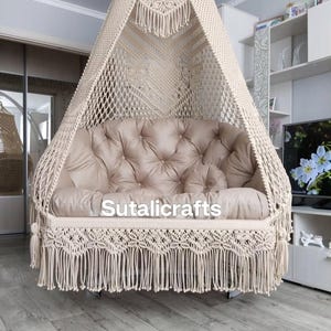 May include: A beige macrame hanging chair with a tufted cushion seat and fringe detailing. The chair is suspended from the ceiling and features the text "Sutalicrafts" on the front. The chair is a bohemian style.