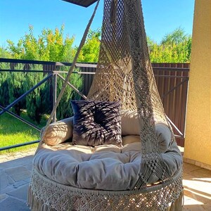 May include: A beige macrame hanging chair with a fringe border and a thick beige cushion. A dark gray pillow is placed on the cushion.