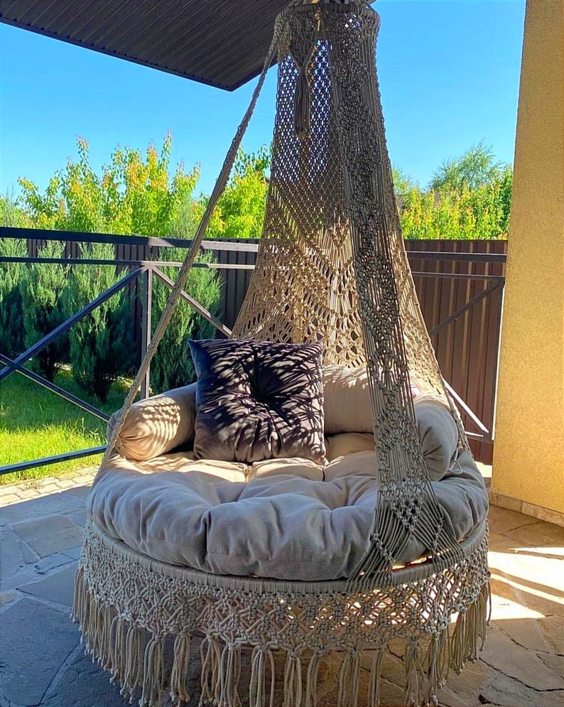 May include: A beige macrame hanging chair with a fringe border and a thick beige cushion. A dark gray pillow is placed on the cushion.